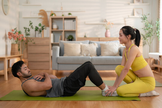 Portrait young Asian couple doing workout together at home, woman supporting partner during sit-ups with care and motivation