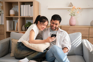 Portrait young Asian couple enjoy watching content on mobile phone together on cozy sofa, laughing with happiness in living room at home
