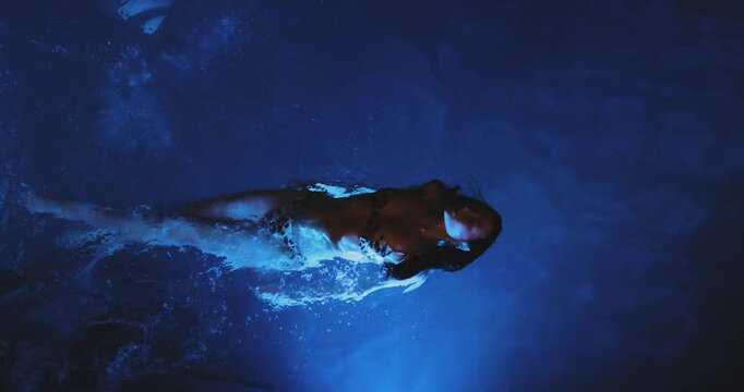 A stunning woman swims gracefully in a glowing blue pool at night, captured in super slow motion from above. The aerial perspective highlights the mesmerizing movement and luminous water. 