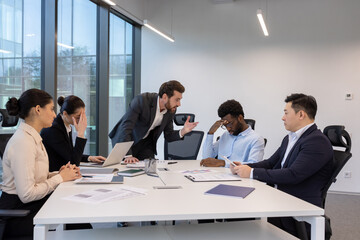 A tense business meeting shows signs of frustration and disagreement among a diverse group of professionals. Some appear worried.