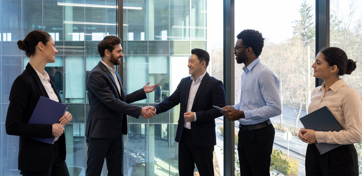 Diverse group of business professionals shaking hands in a modern office, symbolizing agreement or partnership, smiling at each other.