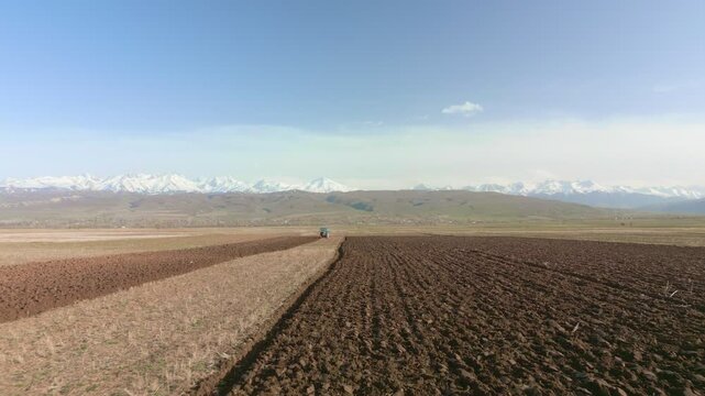 Blue Tractors sowing on agricultural field in spring, readying the field for planting. Powerful tractor turns the soil against the backdrop of high mountains. Issyk-kul Region valley in Kyrgyzstan