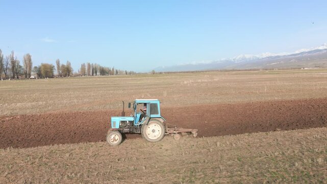 Blue Tractors sowing on agricultural field in spring, readying the field for planting. Powerful tractor turns the soil against the backdrop of high mountains. Issyk-kul Region valley in Kyrgyzstan