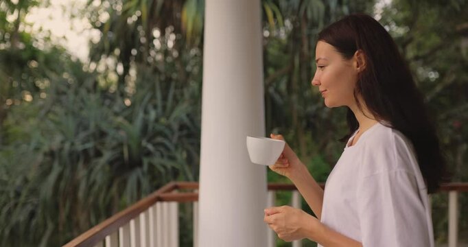 A serene woman in an oversized white shirt sips coffee on a scenic balcony, surrounded by lush tropical greenery. She embraces the peaceful morning atmosphere, enjoying relaxation and mindfulness.