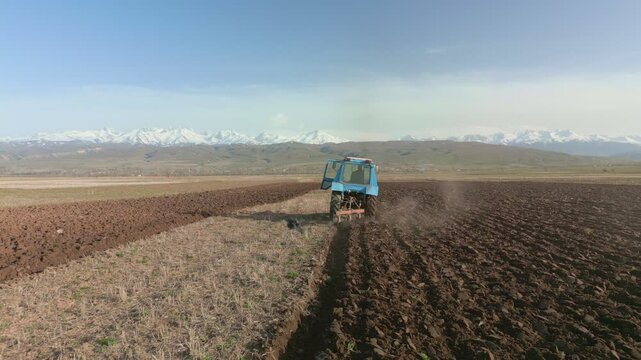 Blue Tractors sowing on agricultural field in spring, readying the field for planting. Powerful tractor turns the soil against the backdrop of high mountains. Issyk-kul Region valley in Kyrgyzstan