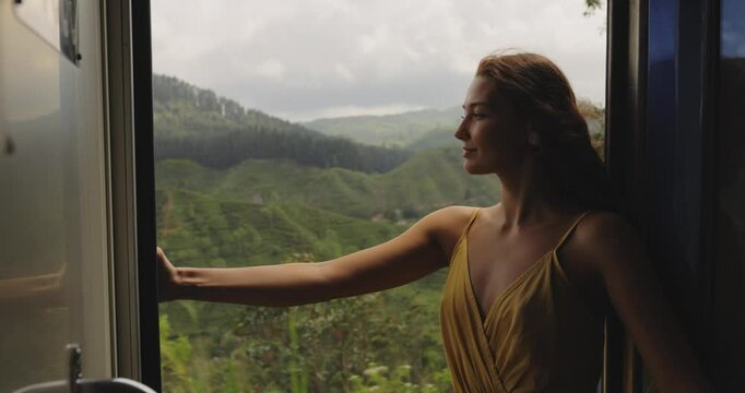 A young woman leans out of a train window, enjoying the fresh air and stunning views of green hills during the train journey from Nuwara Eliya to Kandy, Sri Lanka. Ideal for travel, adventure, and