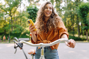 Smiling woman with phone riding bicycle on sunny street in park. Young woman enjoying sunny day and...