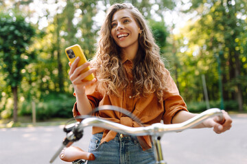 Smiling woman with phone riding bicycle on sunny street in park. Young woman enjoying sunny day and having fun outdoors. Weekend and blogging concept.