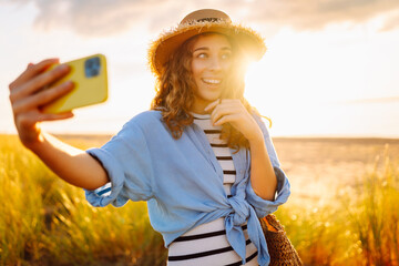 Cheerful woman in hat and modern clothes and phone enjoys sunset on beach. Beautiful tourist takes...