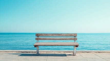 A weathered wooden bench sits on a paved walkway overlooking a calm ocean under a clear blue sky.
