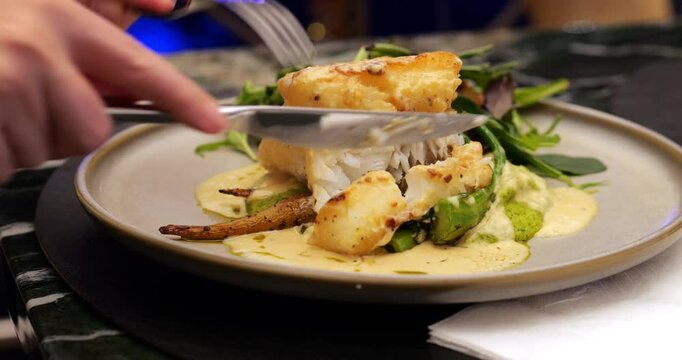 Tourist woman start to eat fish meal, cut piece of fillet by kitchen knife. Evening restaurant in Singapore serving gourmet cod fish with crispy golden crust