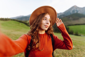 Portrait of woman in hat taking selfie on green mountain hill background. Young female traveler blogging and enjoying sunset landscape. Adventure, blogging and freedom concept.