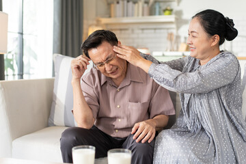 An elderly husband suffering from a headache while his caring wife gently massages and comforts him on the sofa at home. A warm, intimate moment of love, care, and support in senior life