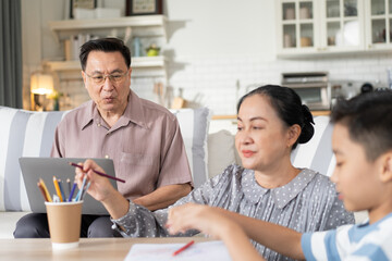 A grandmother watching her grandchild do homework at the table while the grandfather works on a laptop on the sofa in a cozy living room. A warm, multigenerational family moment