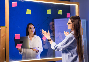 Bright office brainstorming. A young woman collaborates with a laptop, using colorful sticky notes on a glass board to outline ideas in an office.