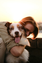Young man hugging his Australian Shepherd dog at sunset. The dog smiles joyfully while warm backlight highlights the bond between them in a serene, golden hour setting.