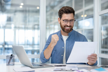 A happy man is celebrating a win at the office, excited with his paperwork, and clenching his fist.