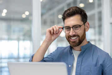 A smiling businessman wearing glasses looks at his laptop. He is in a modern office, possibly at a...
