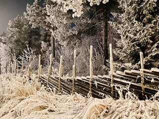 A rustic Norwegian split-rail wooden fence captured during a snowy winter night, surrounded by frost-covered trees and frozen vegetation