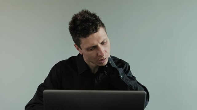 Man feeling neck pain while working on laptop. Uncomfortable working position causing neck strain. Office worker in a black shirt on a gray background