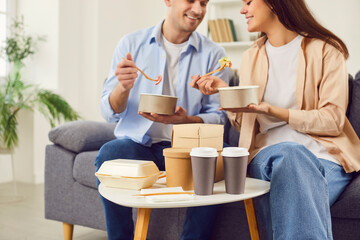 Smiling couple sitting on sofa having fun chatting and enjoying delivered food. Cropped image of...
