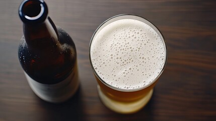 Top-down view of a beer bottle next to a filled glass with a foamy head, placed on a dark wooden surface. 