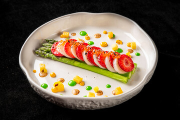 A vibrant dish featuring fresh asparagus topped with sliced strawberries, garnished with colorful dots of sauce and diced mango, creating a visually appealing and flavorful appetizer.