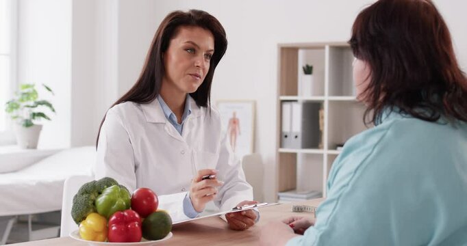 Pretty friendly nutritionist woman talking with her fat female patient and making notes in notepad about diet plan for weight loss sitting in office. Healthy food and nutrition concept. 4k video.