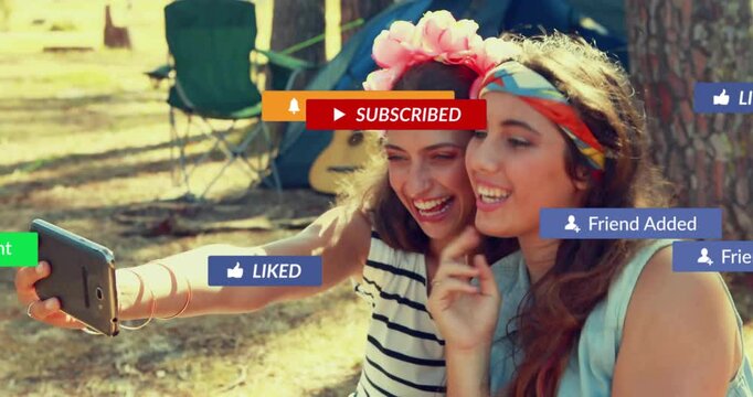 Two friends taking selfie at campsite with tent, using travel app overlay with floating phone icons