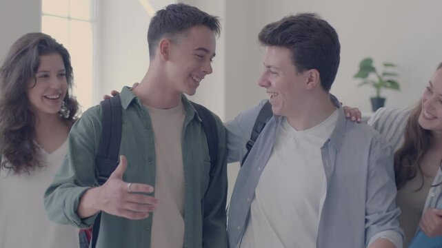Several high school pals meet in the classroom. Group of smiling, cheerful teenage student boys and girls with backpacks greet each other before class. Two happy friends say hello and hug each other