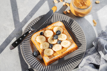Peanut butter toast with banana and blueberry on a plate on a light background with morning shadow.