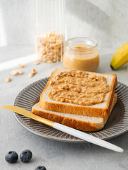 Peanut butter toast on a plate on a light background with banana, blueberry and morning shadow
