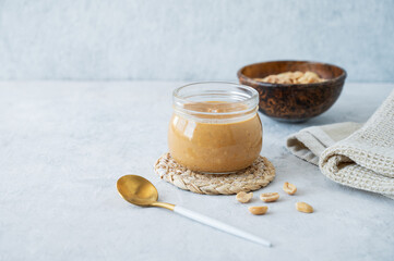 Peanut butter in glass jar on light background with salted nuts and napkin.