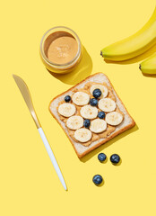 Peanut butter toast with banana, blueberry, nuts and knife on a yellow background.