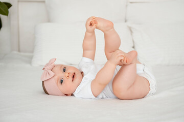 A baby girl is smiling at home on a white bed with a bow on her head, a happy healthy child of six months is playing with her legs on the back