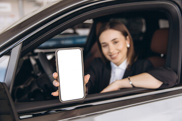 Businesswoman showing smartphone with blank screen sitting in new car at dealership