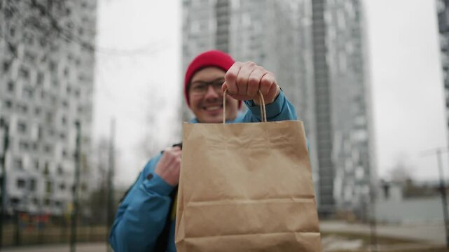 Professional delivery worker wearing red hat and glasses, handing brown paper bag to camera with blurred urban setting in background, representing modern food delivery service