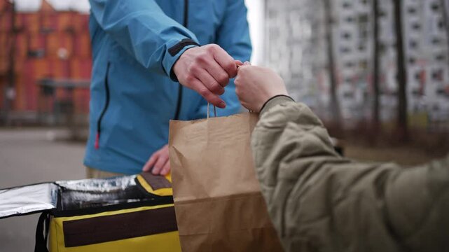 Urban bicycle messenger delivering package from insulated yellow backpack, handing brown paper bag to recipient on busy street, representing quick urban delivery