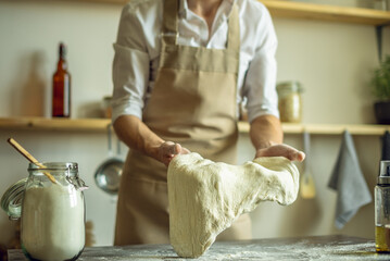 A baker in an apron kneads the dough with his hands and sprinkles it with flour to bake delicious homemade bread.