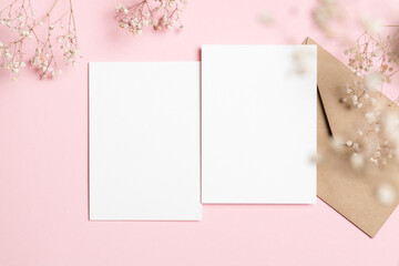 Two vertical white blank card mockups with kraft envelope and baby breath flowers on pastel pink background, ideal for wedding invitation design, flat lay