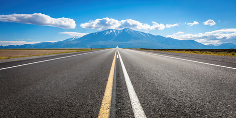 Empty asphalt road with clear lane markings stretches toward distant mountain under bright blue sky with scattered clouds, evoking sense of freedom and adventure