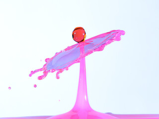 A vibrant pink water splash with a dark red droplet colliding at its peak, captured mid-air against...