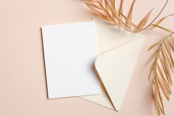 Blank wedding invitation or greeting card mockup on a beige background with envelope and dried palm...