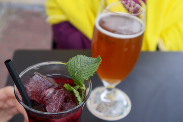 
Two glasses are visible on a dark table: one with a dark red drink, ice cubes, a black straw and a mint leaf, and the other with a light, frothy drink, probably beer.