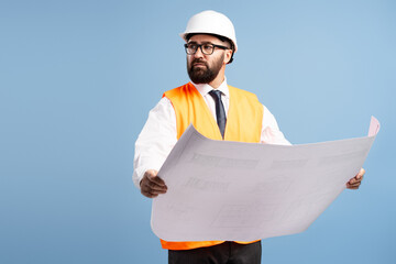 Confident engineer wearing safety helmet and vest reviewing blueprints on blue background