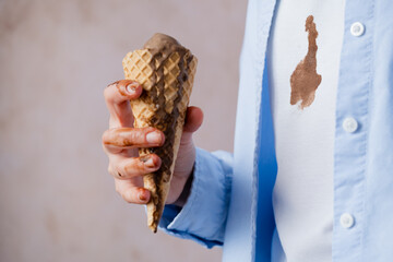 A person holding chocolate ice cream cone, spilled ice cream on white clothes. Ice cream flowing from the cone onto the hand. Concept of daily food stains on clothes