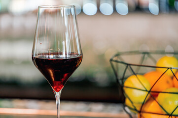 A close-up of a glass of red wine with a shimmering surface, placed beside a wire fruit basket filled with bright oranges, set against a softly blurred background.