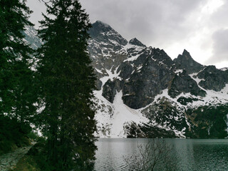 Tall evergreens line a stone path beside Morskie Oko lake, with dramatic snow-covered Tatra...