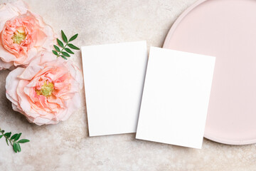 Two white blank wedding cards mockup on a textured surface with roses flowers and green leaves. Elegant flat lay, perfect for invitations or greeting cards design