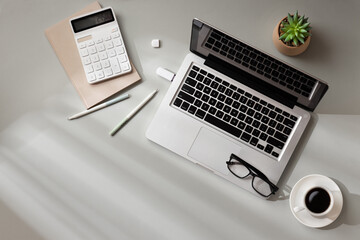 A workspace with a laptop, calculator, notebook, plant, glasses, pens, USB and coffee cup on a light gray surface with sunlight from the window. Top view.